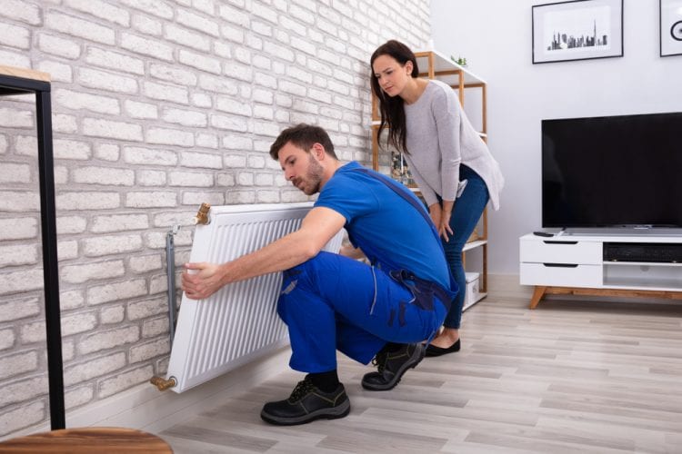 Joven electricista instala un radiador justo a la pared de ladrillos en casa de una mujer Joven electricista instala un radiador justo a la pared de ladrillos en casa de una mujer